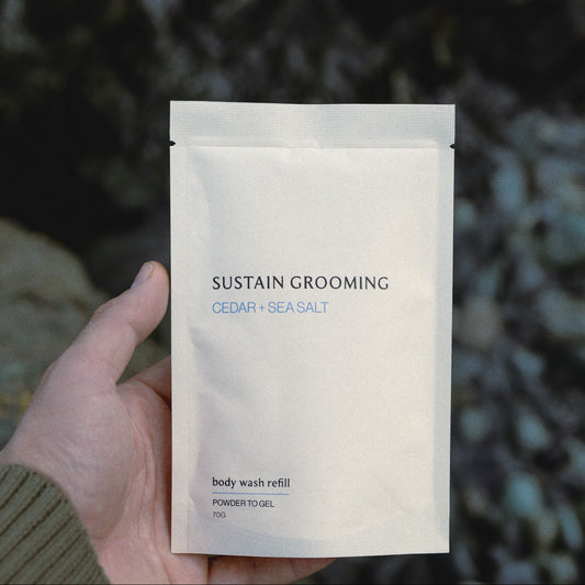 Hand holding a 'Sustain Grooming' body wash refill packet against a natural background.