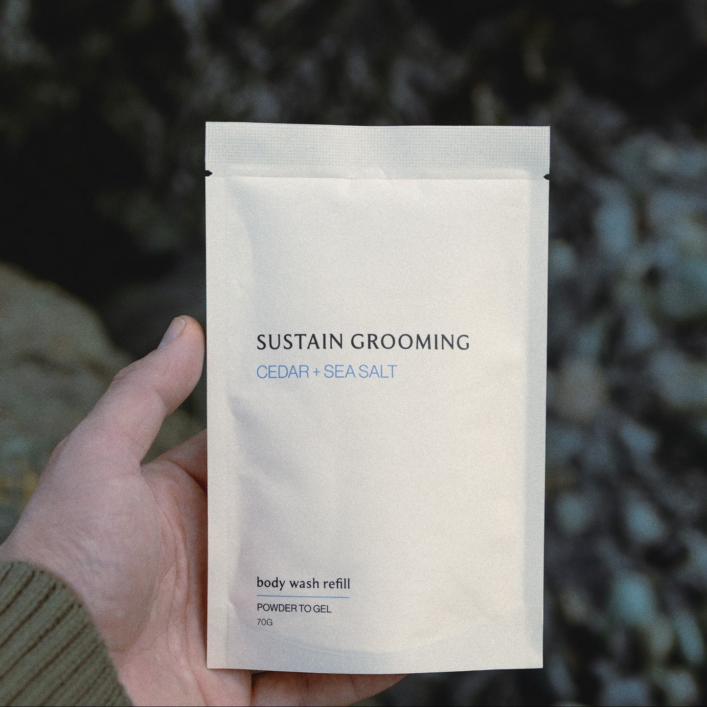Hand holding a 'Sustain Grooming' body wash refill packet against a natural background.