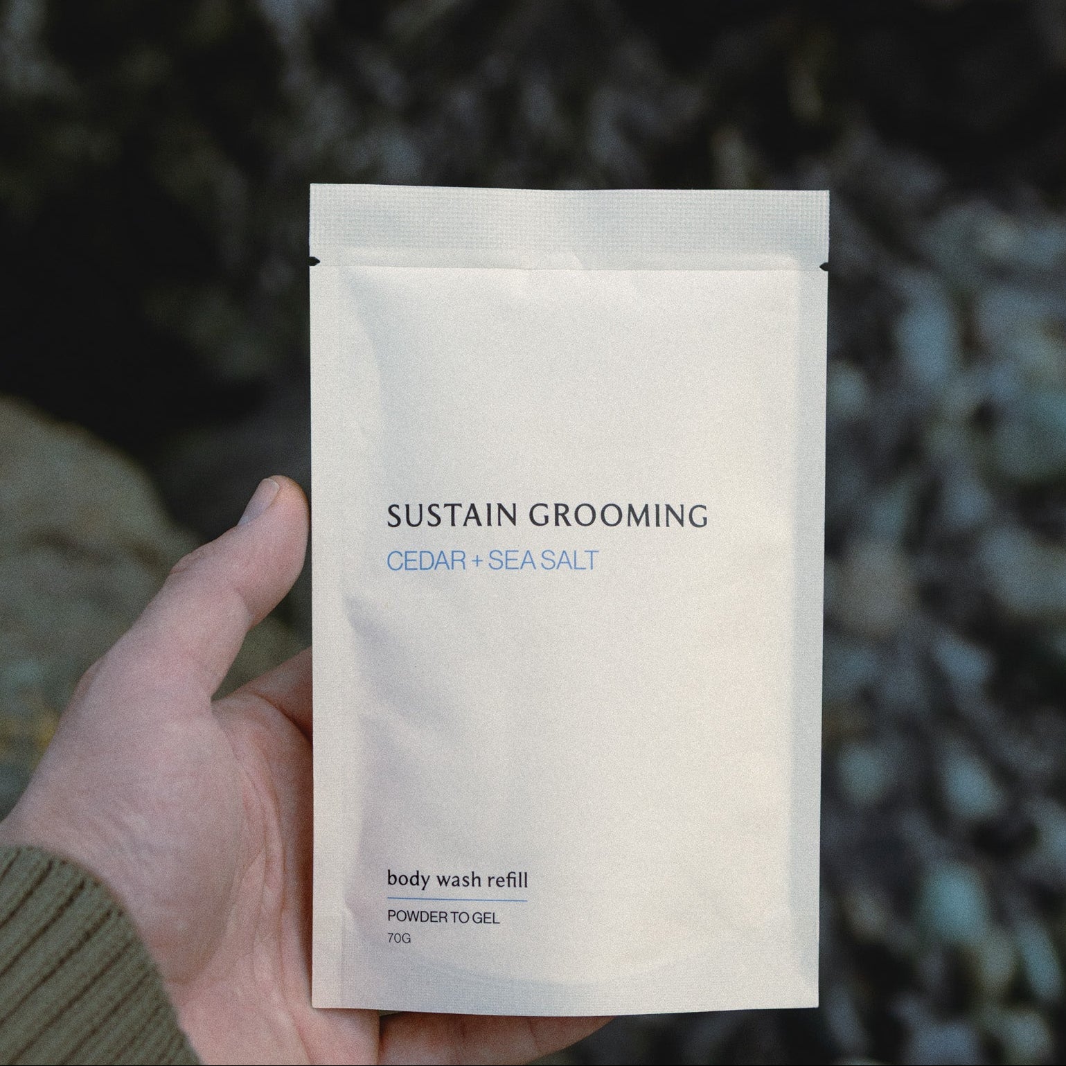 Hand holding a 'Sustain Grooming' body wash refill packet against a natural background.