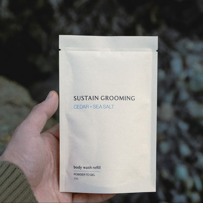 Hand holding a 'Sustain Grooming' body wash refill packet against a natural background.